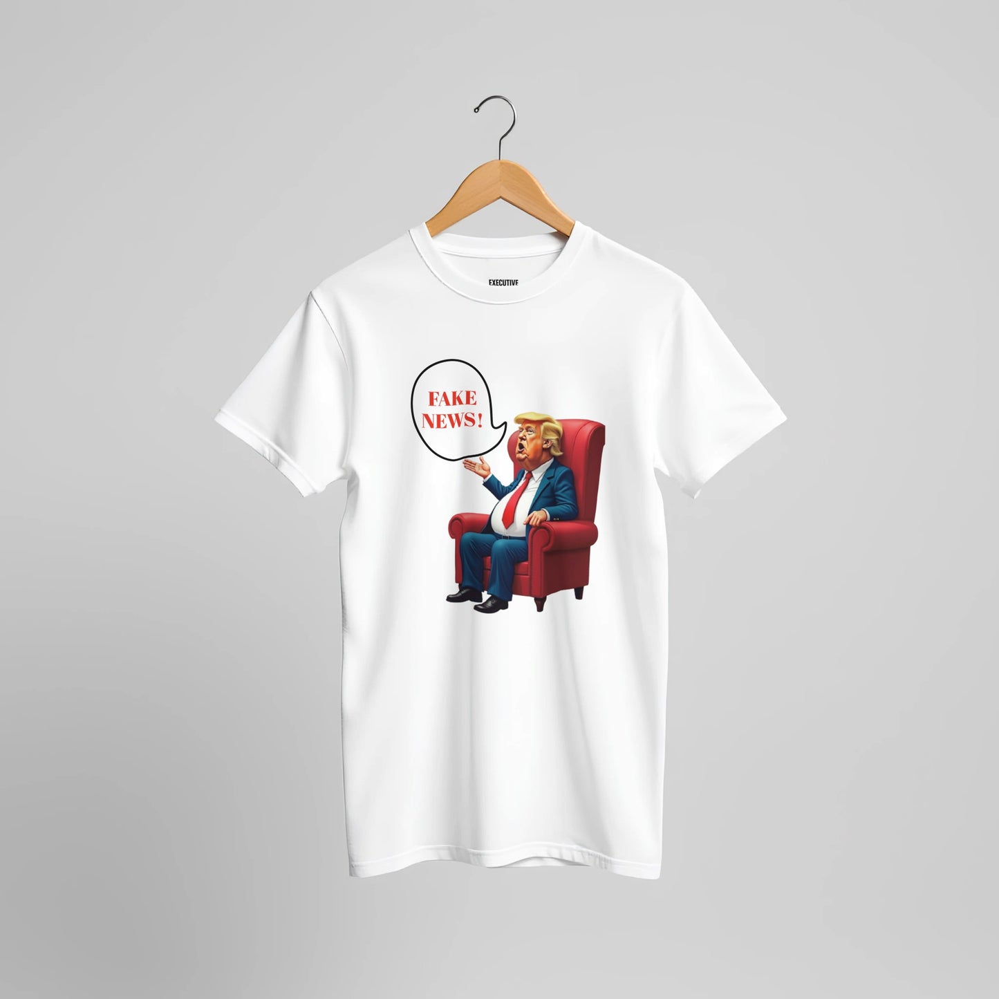 White t-shirt with a colourful cartoon-style caricature of a suited, orange-tinted man sitting in a red armchair, raising one hand and shouting ‘FAKE NEWS!’ in a speech bubble