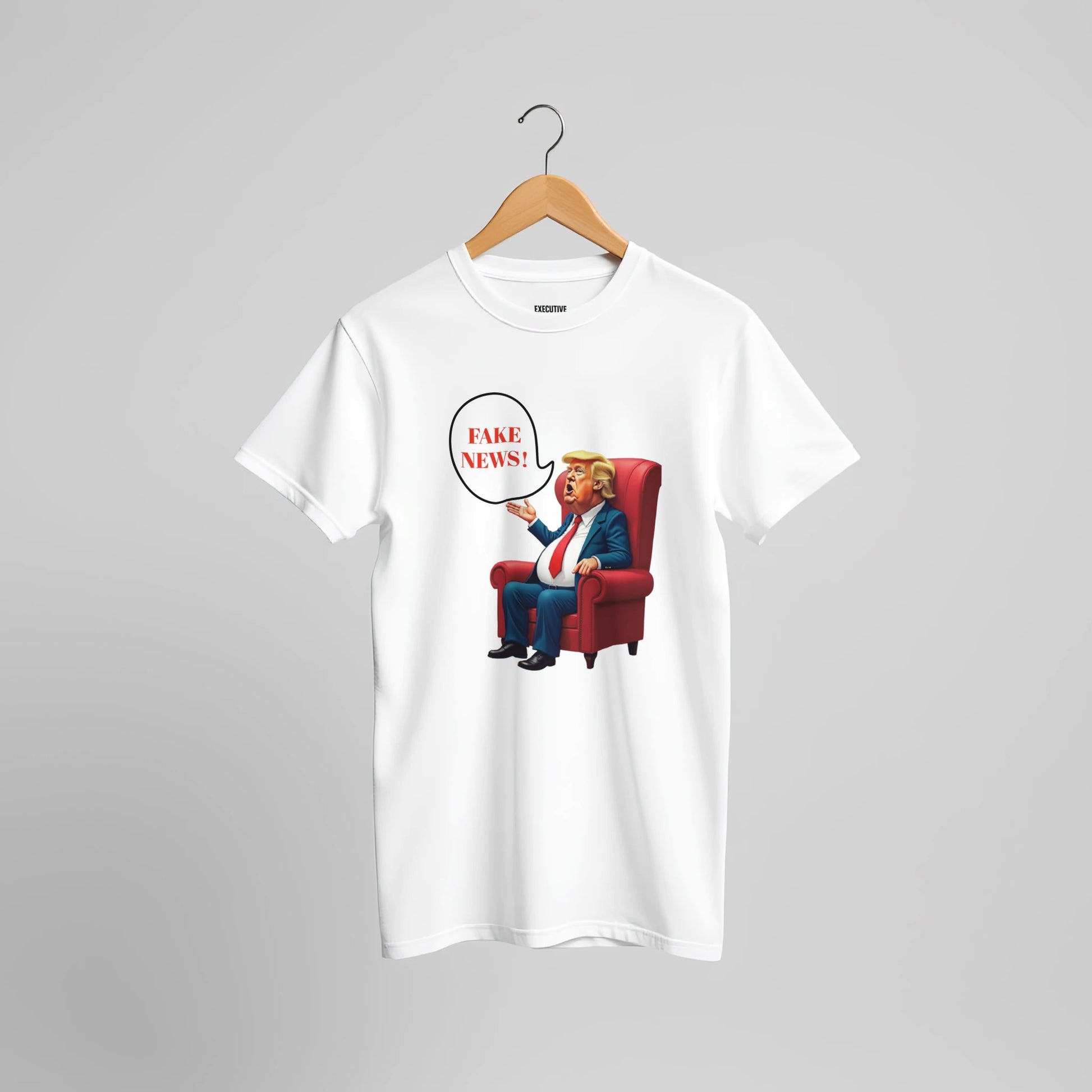 White t-shirt with a colourful cartoon-style caricature of a suited, orange-tinted man sitting in a red armchair, raising one hand and shouting ‘FAKE NEWS!’ in a speech bubble