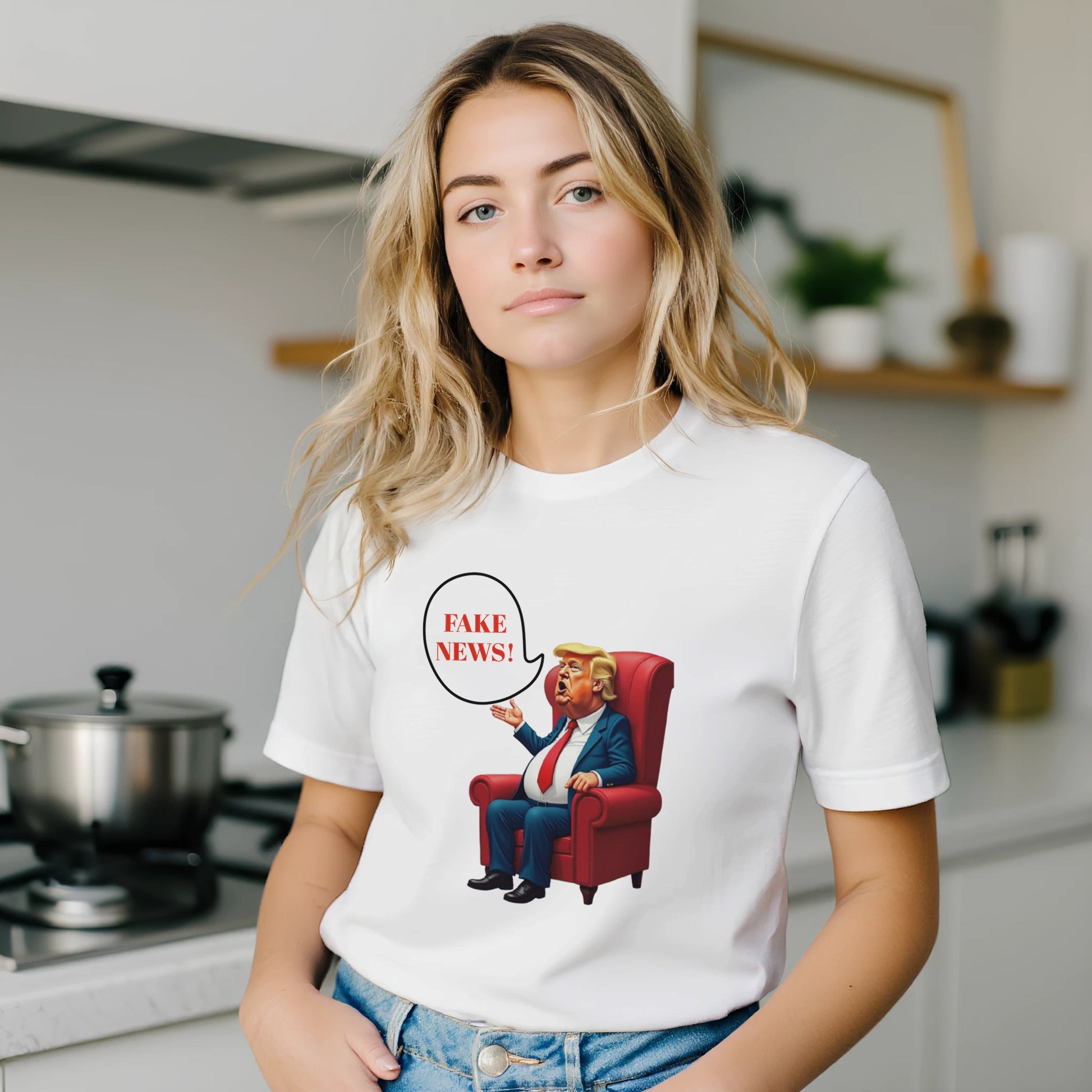 White t-shirt with a colourful cartoon-style caricature of a suited, orange-tinted man sitting in a red armchair, raising one hand and shouting ‘FAKE NEWS!’ in a speech bubble